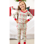 A young girl stands in front of a bed wearing a festive Christmas-themed long-sleeve pajama set. The pajamas feature a light cream background with a repeating pattern of red bows, green holly leaves, and pinecones, with red trim on the collar, cuffs, and ankles.