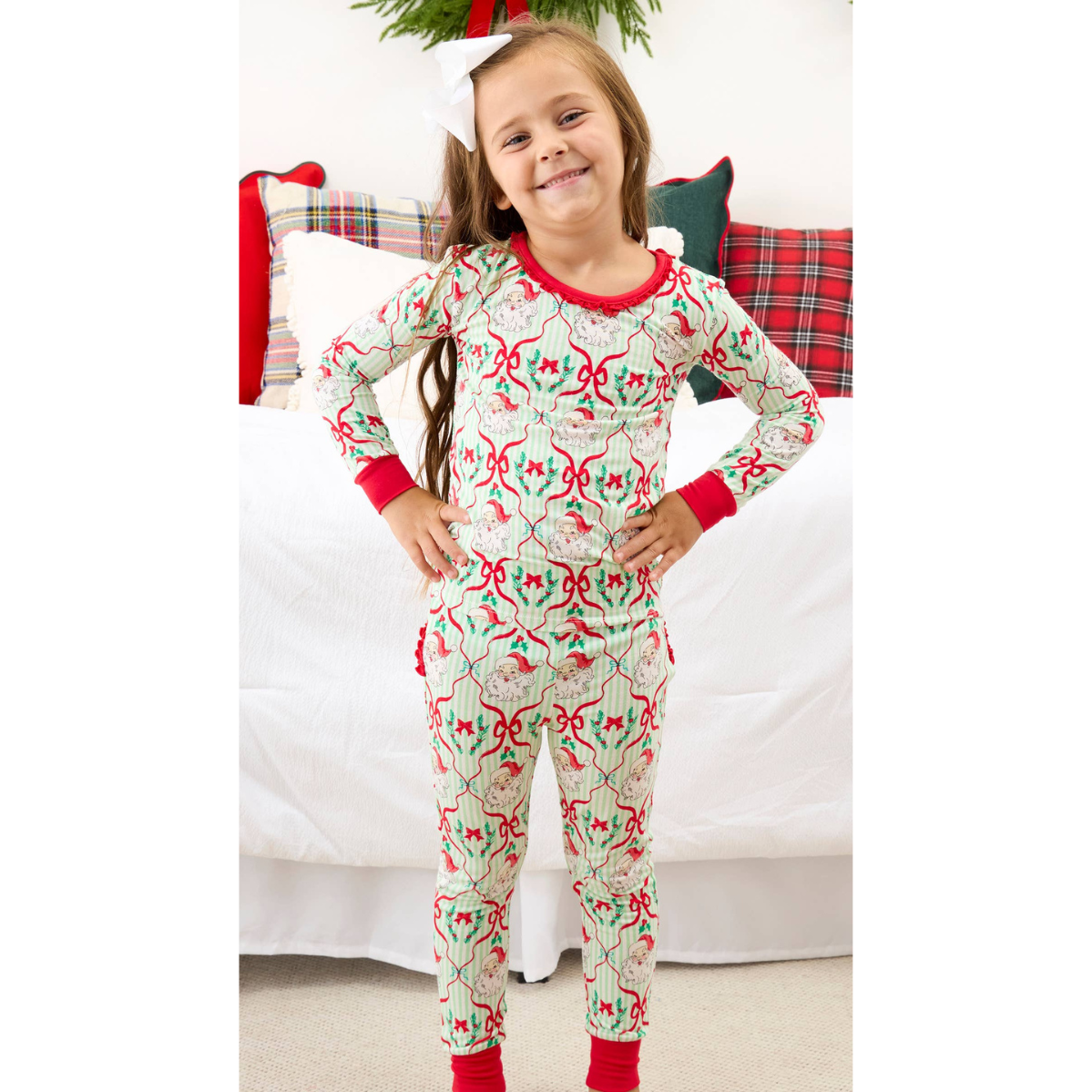 A young girl stands in front of a bed wearing a festive Christmas-themed long-sleeve pajama set. The pajamas feature a light cream background with a repeating pattern of red bows, green holly leaves, and pinecones, with red trim on the collar, cuffs, and ankles.