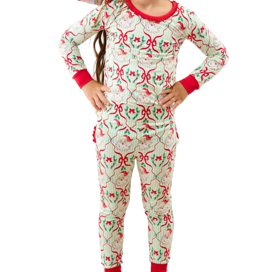 A young girl stands in front of a bed wearing a festive Christmas-themed long-sleeve pajama set. The pajamas feature a light cream background with a repeating pattern of red bows, green holly leaves, and pinecones, with red trim on the collar, cuffs, and ankles.