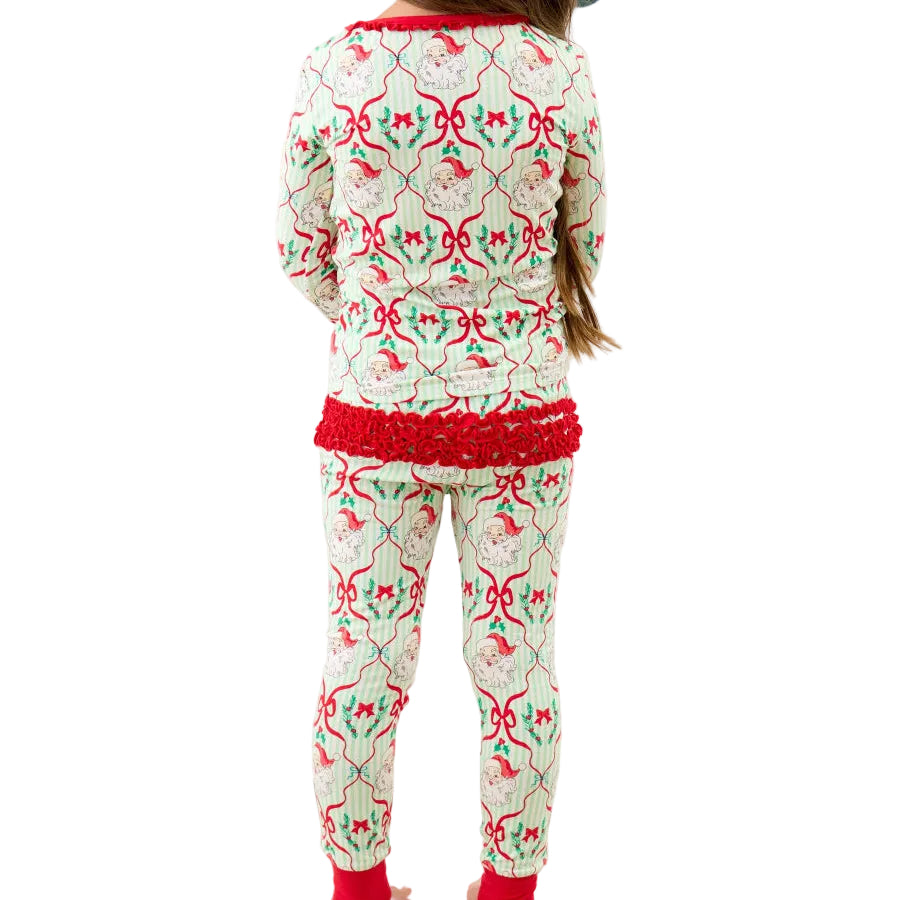 A child viewed from behind wearing a festive Christmas-themed pajama set with a red and green patterned top and matching pants, standing in front of a bed decorated with holiday pillows.