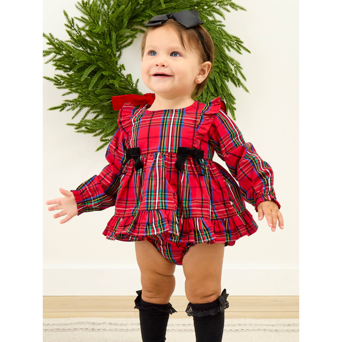 A baby girl wearing a festive red plaid long-sleeve romper dress with black bow accents, black knee socks, and a black bow headband, standing in front of a green wreath.