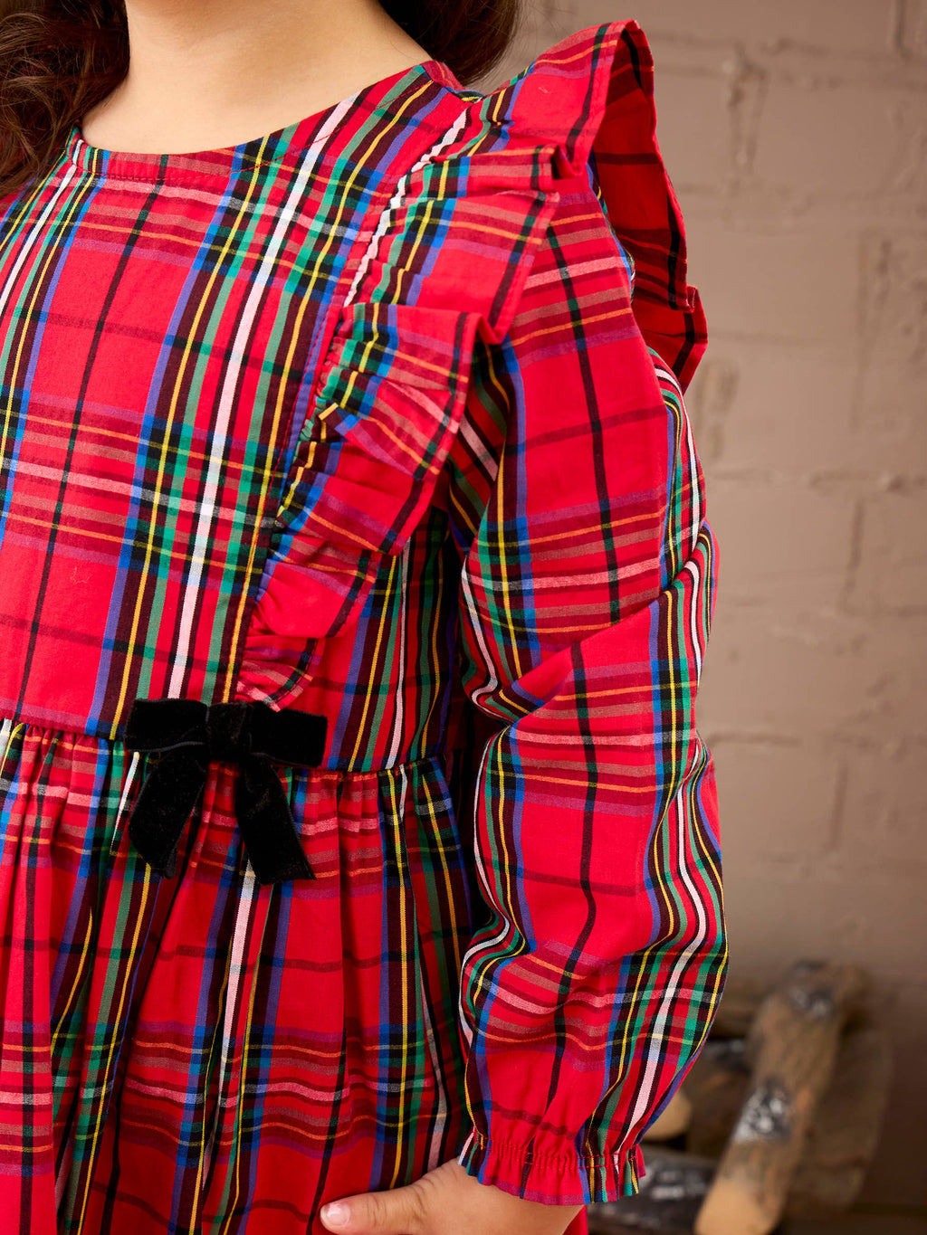 A close-up of a child wearing a bright red plaid dress with multicolored stripes, ruffled sleeves, and a black bow at the waist, standing against a light brick wall.