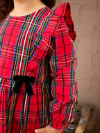 A close-up of a child wearing a bright red plaid dress with multicolored stripes, ruffled sleeves, and a black bow at the waist, standing against a light brick wall.