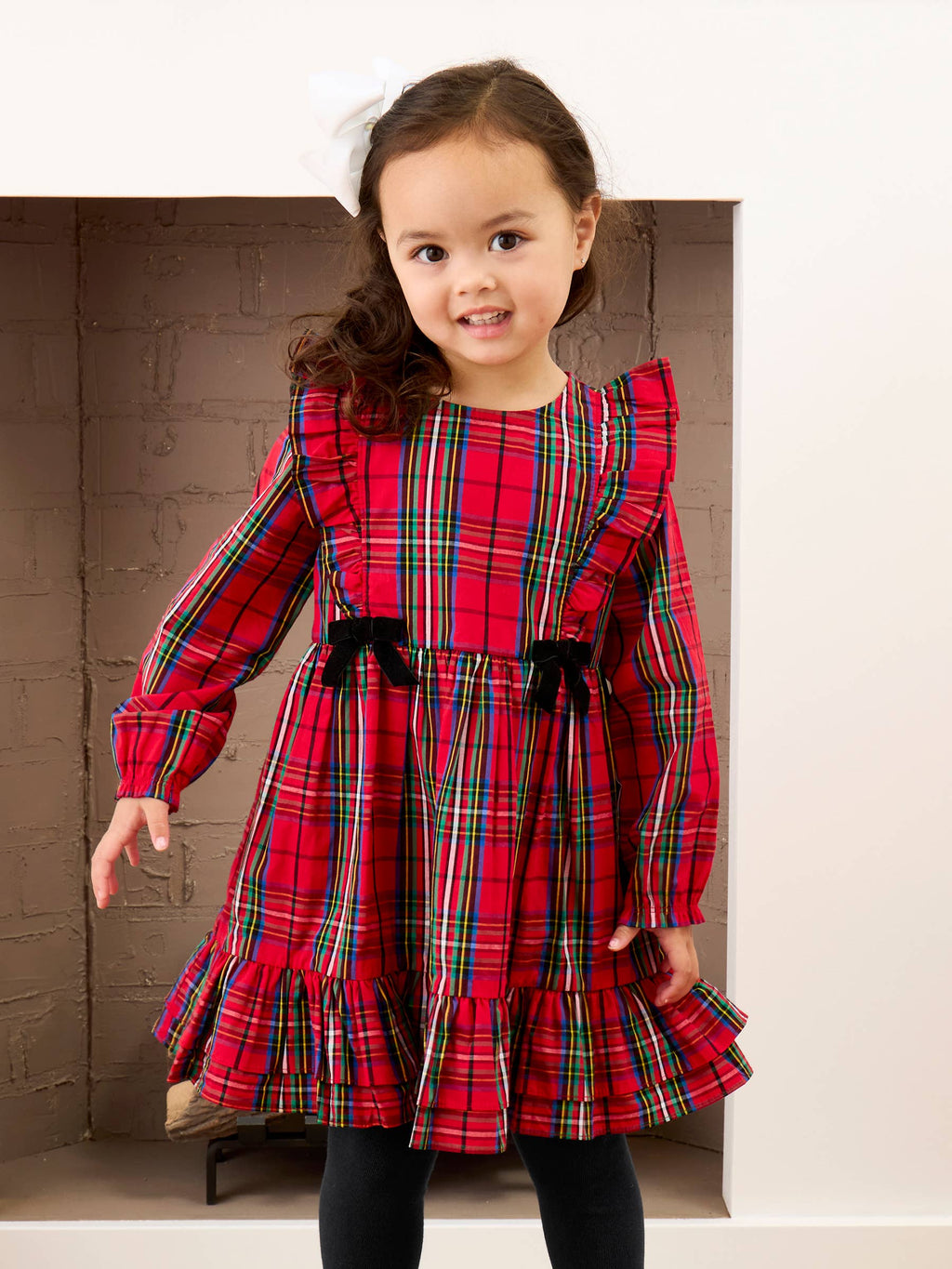 A young girl wearing a red tartan plaid dress with black ribbon bows at the waist, standing in front of a brick fireplace.
