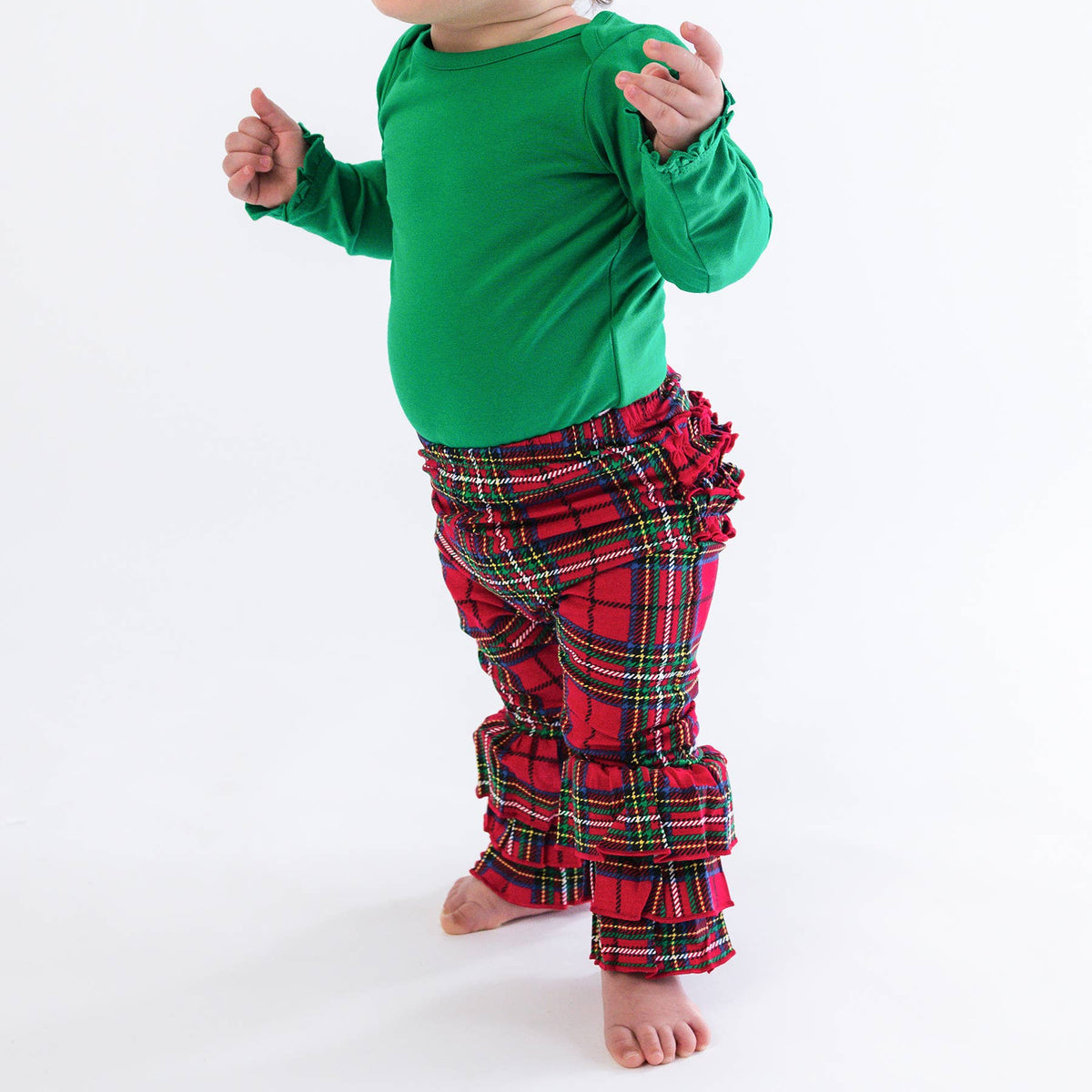 A baby wearing a bright green long-sleeve top and red plaid ruffled pants, standing barefoot on a white studio background. The baby also wears a red bow headband.