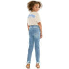 A young girl with curly brown hair is shown from the back, wearing light-wash blue jeans with a raw hem, a cream-colored graphic t-shirt, and brown sandals, set against a plain white studio background.