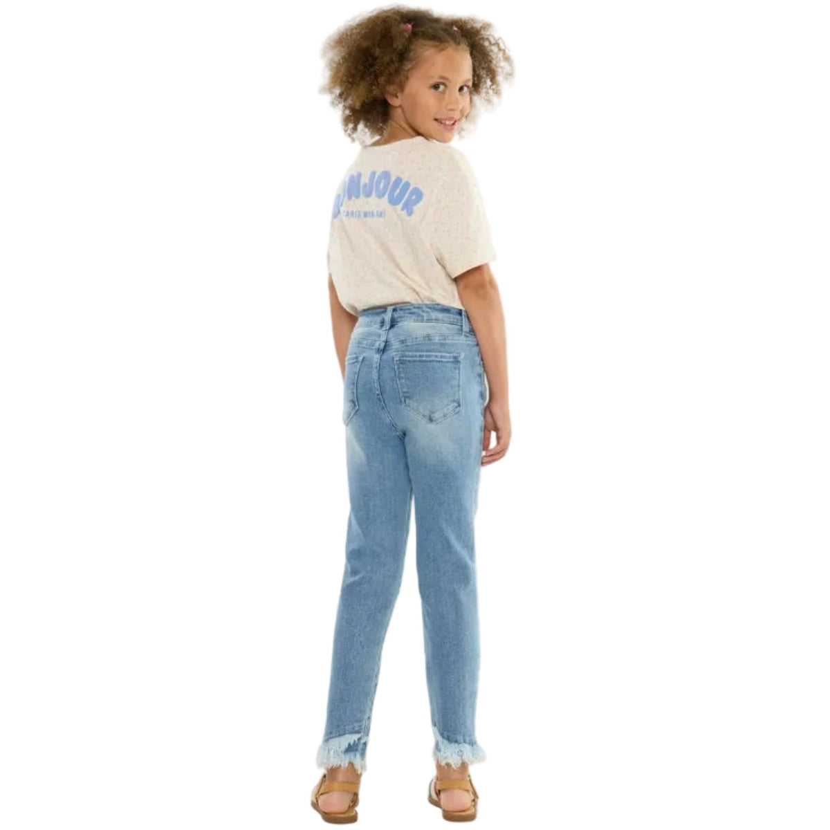 A young girl with curly brown hair is shown from the back, wearing light-wash blue jeans with a raw hem, a cream-colored graphic t-shirt, and brown sandals, set against a plain white studio background.