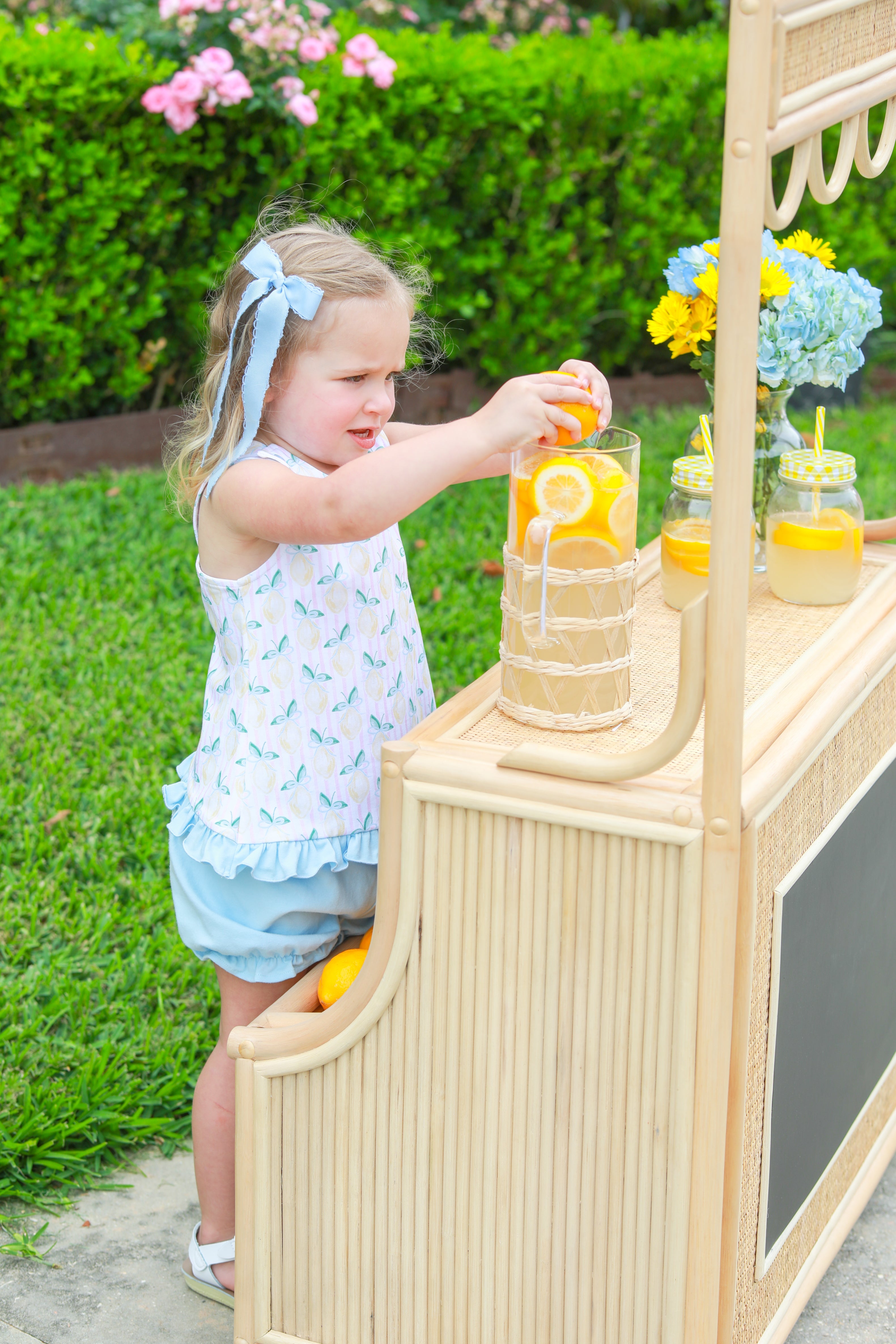 Toddler girl at a lemonade stand wearing a lemon print bloomer set with pink stripe top and blue bloomers.
