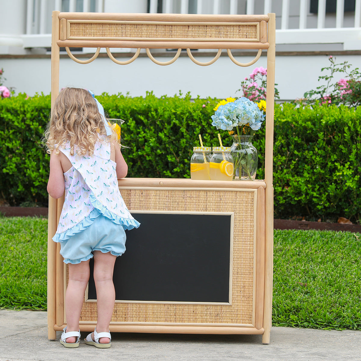 Little girl standing at a lemonade stand wearing a lemon print bloomer set with pink stripe top and blue bloomers.