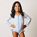 Young girl wearing floral one-piece rash guard swimsuit with long sleeves, zipper front, and bows at the hips.
