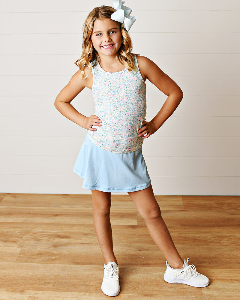 Young girl wearing a floral tank top and light blue skort athleisure outfit with white sneakers.
