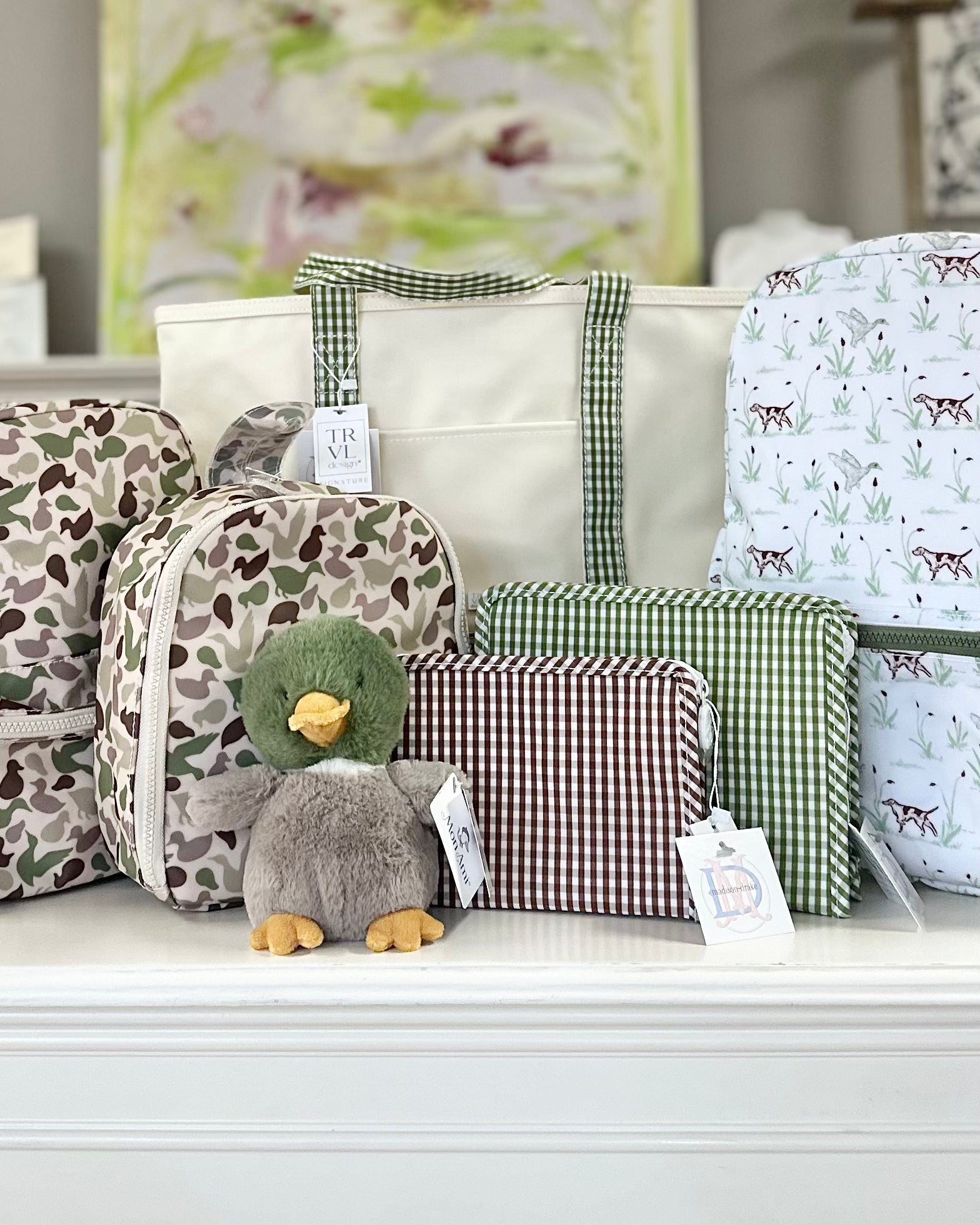 A collection of moss, camouflage, and gingham TRVL Design bags including a canvas tote bag, a backpack, a lunch bag, a pouch, and a small plush duck toy.