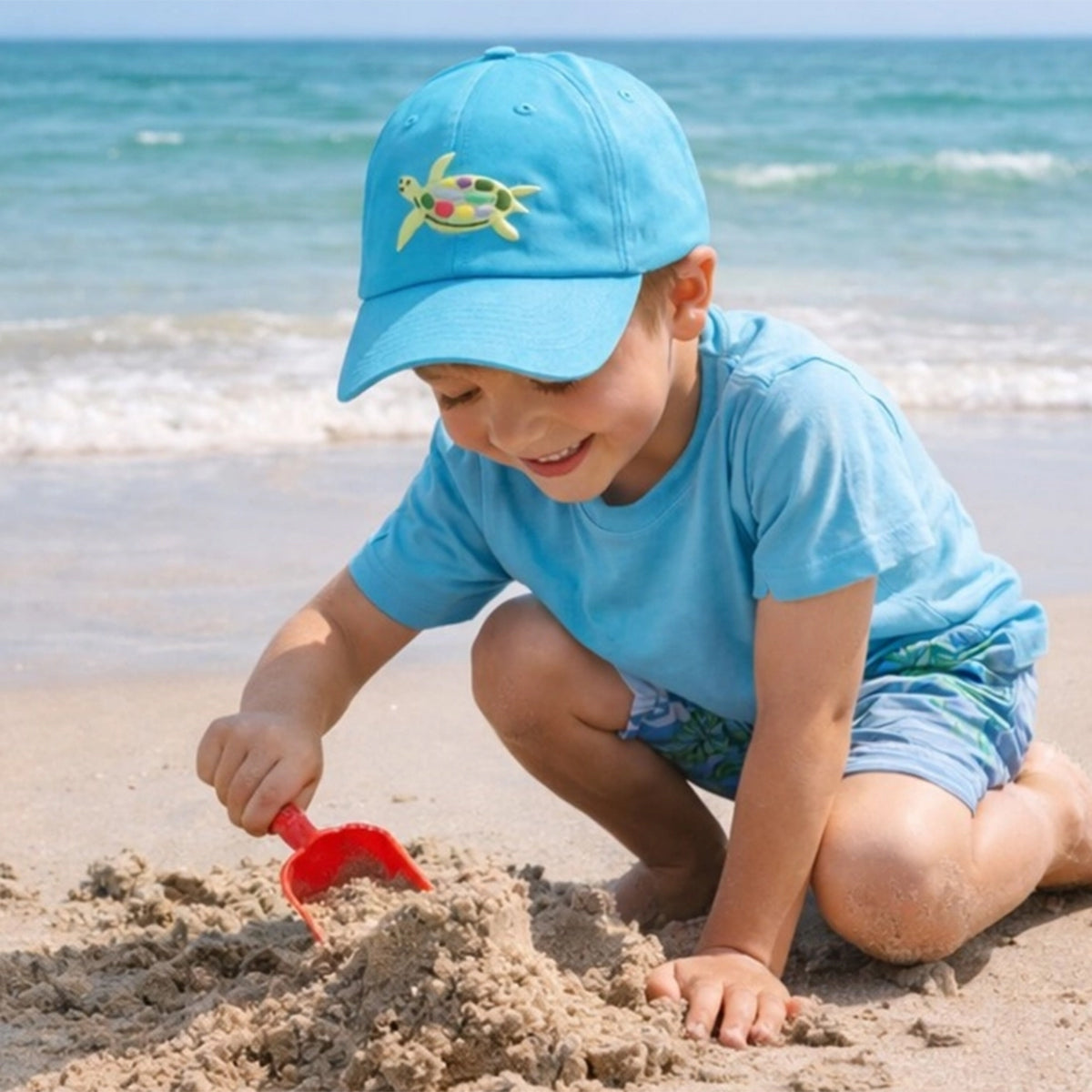 Little boy wearing a turquoise baseball cap with an embroidered sea turtle design playing on the beach.