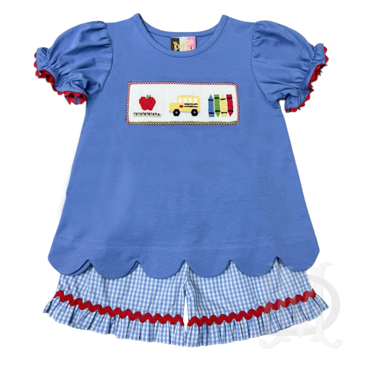 A girl's back to school outfit with a medium blue short sleeve shirt and scalloped hem featuring school-themed smocking with an apple, bus, and crayons. The shorts are blue-and-white gingham check with a ruffled trim and red ric-rac.