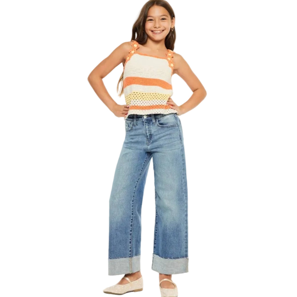 A young girl modeling a knit striped tank top with orange shoulder straps and wide-leg blue jeans with cuffed hems, standing against a plain white studio background.