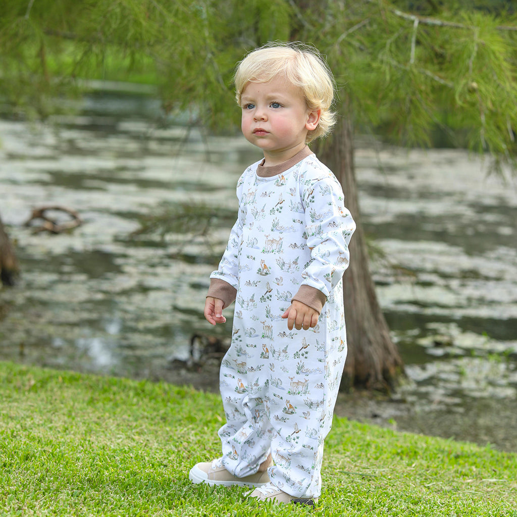 Child wearing a white onesie with animal prints standing on grass near a body of water.