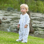 Child wearing a white onesie with animal prints standing on grass near a body of water.